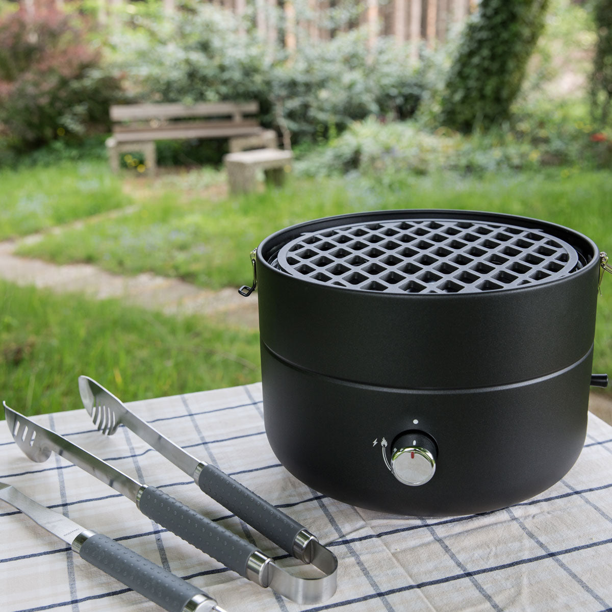 Der Grill-to-Go mit Gusseisen Grillrost steht auf einem Tisch mit einer Grillzange daneben