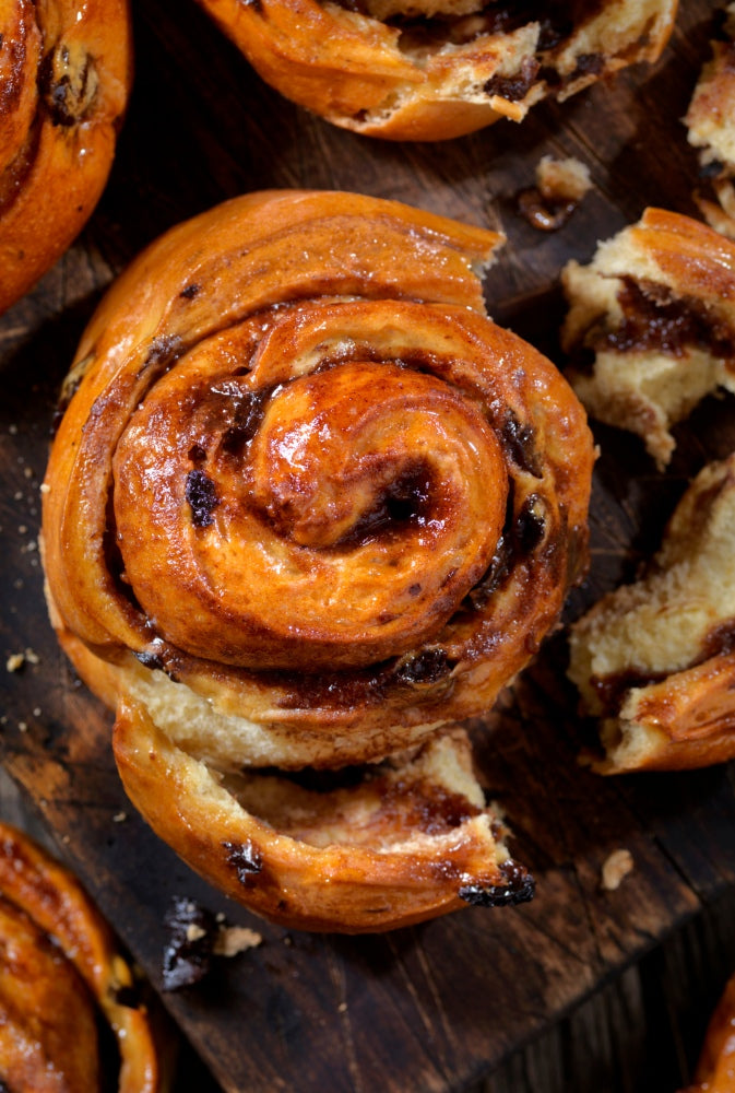 Die Zimtschnecken aus dem Dutch Oven liegen auf einem Holzbrett bereit verzehrt zu werden. Von einer Zimtschnecke ist ein Stück abgerissen, sodass man die saftige Füllung sehen kann.