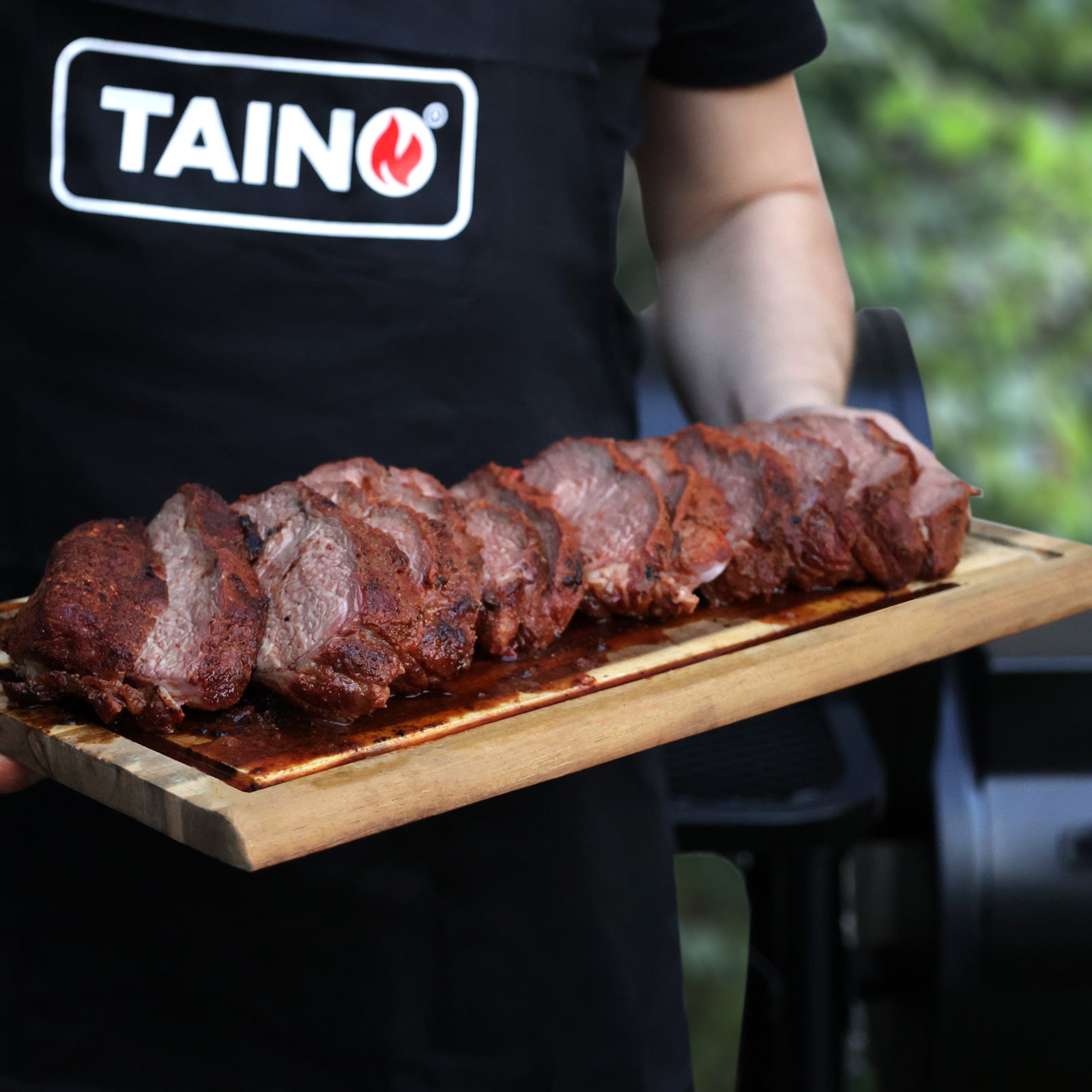 Eine Person mit einer Schürze mit TAINO Logo hält ein Holzbrett  mit gesmoktem Fleisch 
